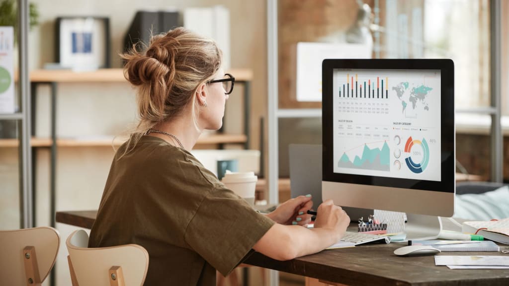 A woman in glasses sits at a desk, looking at a computer screen displaying charts and graphs. She's holding a pen and surrounded by papers and office supplies.